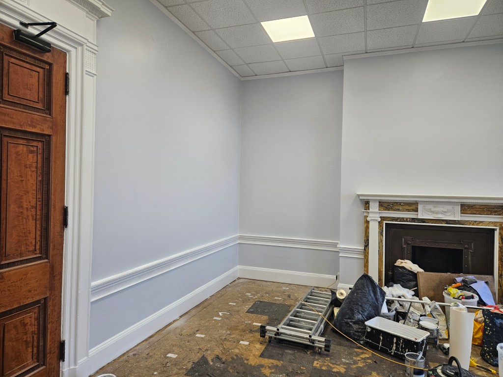 Interior view of a room with light gray walls, a wooden door on the left, and a fireplace with decorative trim. The floor is cluttered with renovation materials including paint cans and debris.