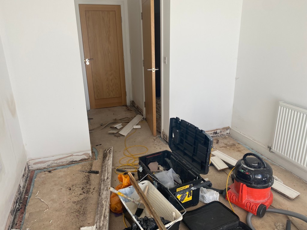 An unfinished room with bare flooring and construction debris, including tools and a vacuum cleaner, alongside an open doorway leading to another room.
